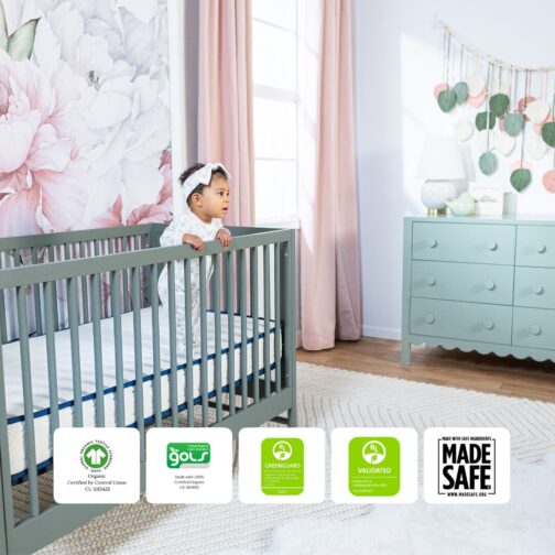 Baby standing in a crib on the Emily Organic Crib Mattress with GOTS, GOLS, GREENGUARD Gold, and MadeSafe certifications displayed.