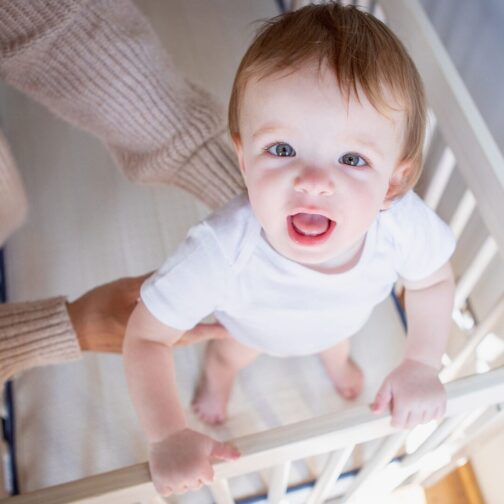 Baby smiling while standing in a crib featuring the Emily Organic Crib Mattress with safe, durable two-sided construction.